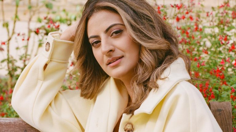 We see a photo Harpz looking at the camera and wearing a pale yellow jacket. She's sitting on a bench with red flowers in the background.