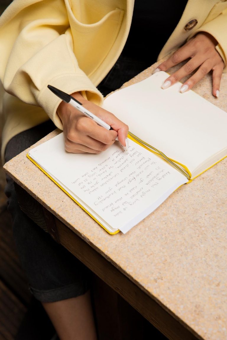 We see a close up shot of Harpz sitting at a table, writing her letter in a yellow notebook.